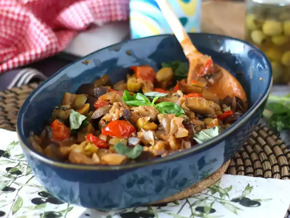 Un plat de caponata recette, un m&eacute;lange color&eacute; d'aubergines, tomates et herbes fra&icirc;ches, servi dans un bol bleu avec une cuill&egrave;re en bois.