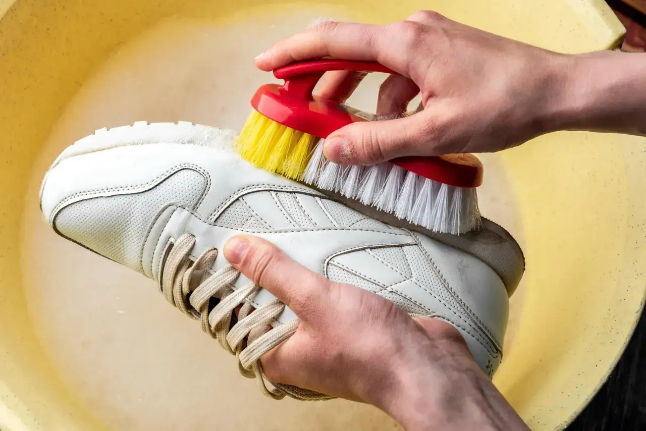 Mani che lavano una Superga bianca con una spazzola rossa e gialla in una bacinella gialla.