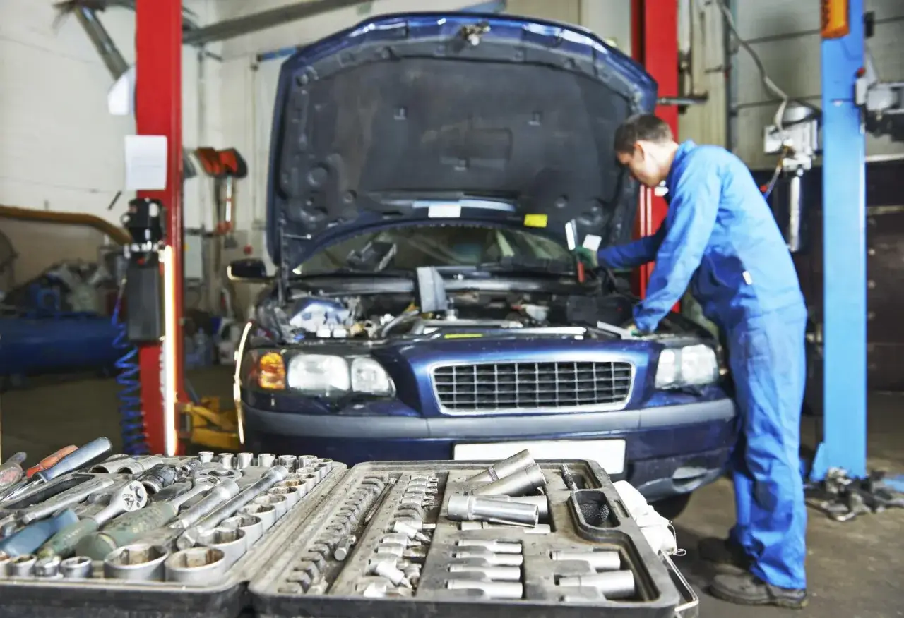 Mechanik w niebieskim kombinezonie naprawia silnik samochodu. Obok leży skrzynka z narzędziami, gotowa do rozwiązania problemu szarpania przy puszczeniu gazu.