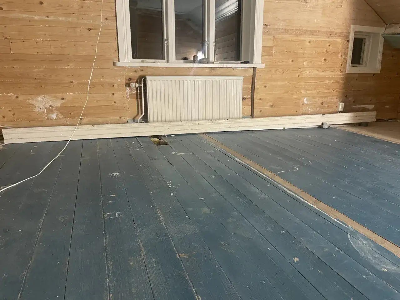 Ett rum under renovering med träväggar och ett slitet, blåmålat **luta golv**. En vit list ligger längs väggen.