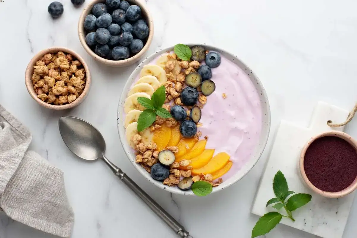 Kolorowa miska z jogurtem, bananami, brzoskwiniami i jagodami to idealne co na śniadanie. Obok granola i łyżka.
