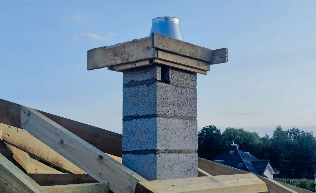 Zakończenie komina systemowego z metalową nasadką, otoczone drewnianą konstrukcją dachu.