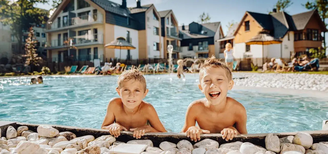Dwóch chłopców cieszy się kąpielą w basenie przy hotelu Karpacz w Karpaczu. W tle widać nowoczesne budynki i leżaki.