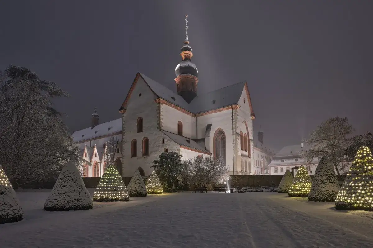 Ein Kloster im Schnee, festlich beleuchtet. Die Bäume sind mit Lichterketten geschmückt, als ob man Weihnachten im Kloster verbringen würde.