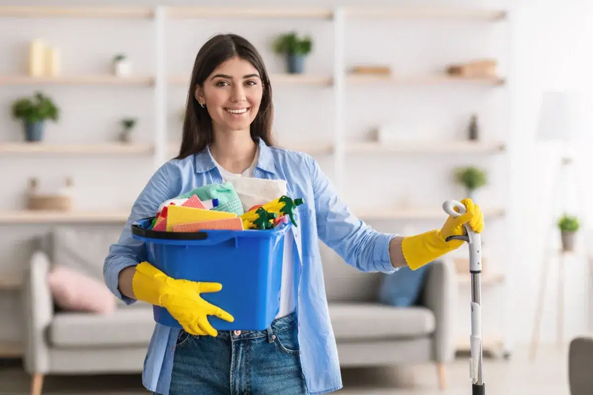 Mujer sonriente con guantes amarillos, lista para limpiar la casa, lleva un cubo azul con productos de limpieza y un mocho.