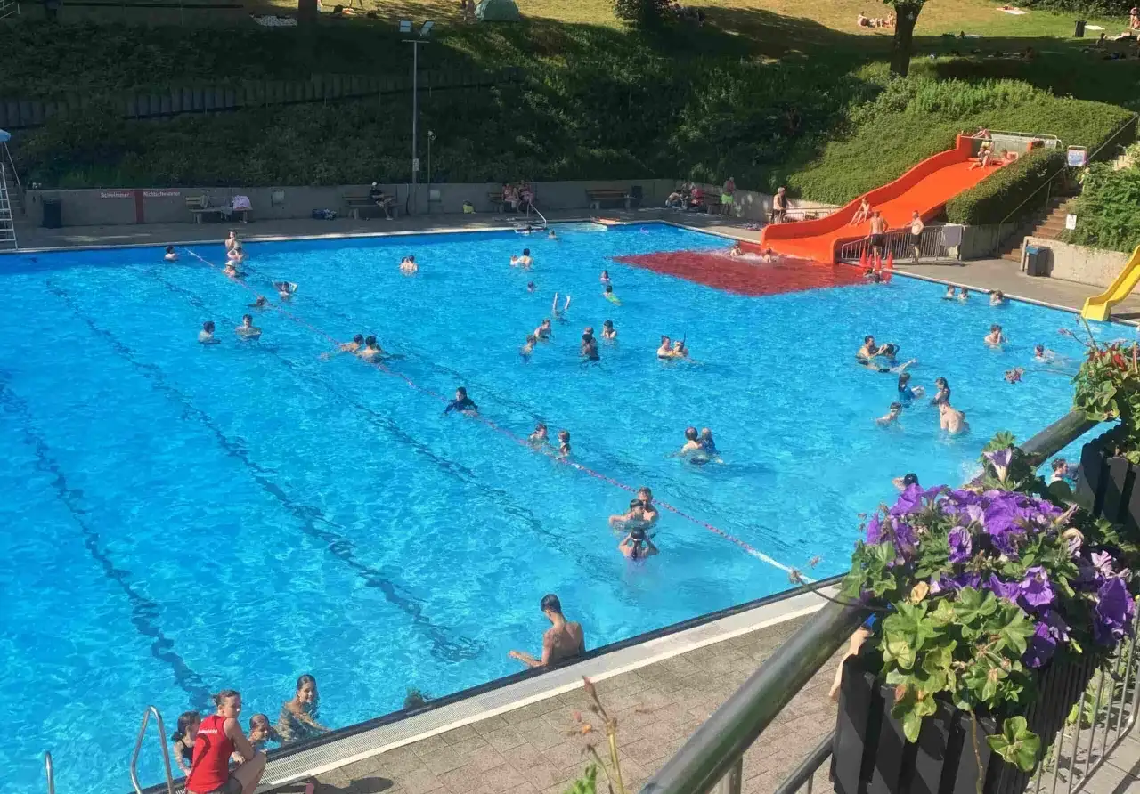 Sonniges Treiben im Freibad Neuenhof: Kinder und Erwachsene planschen im blauen Wasser, rutschen und entspannen am Beckenrand.