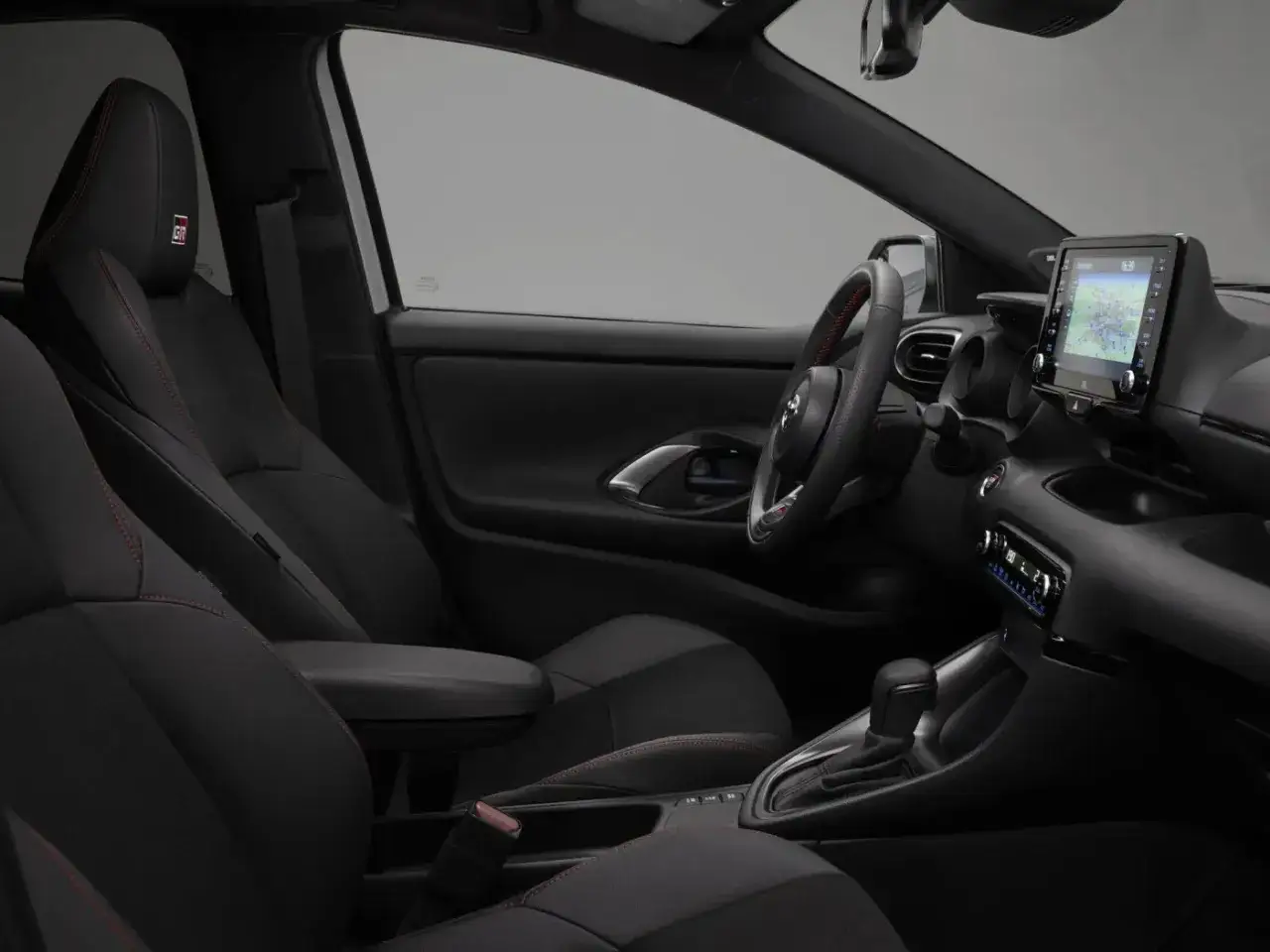 Wnętrze sportowego Toyota Yaris z czarnymi fotelami i czerwonymi przeszyciami. Kierownica i ekran multimedialny widoczne.