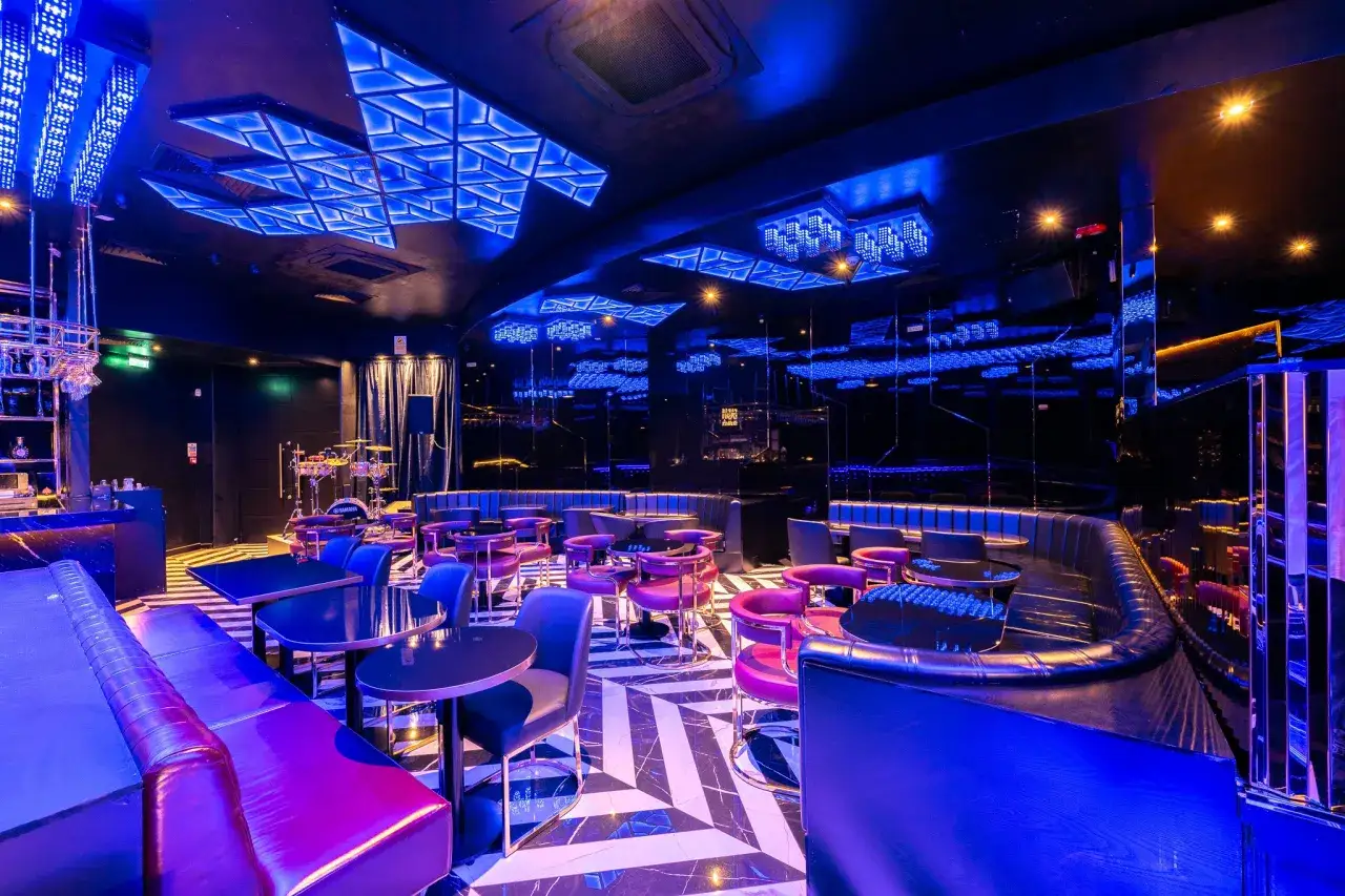 Interni di un club di Londra con divani in pelle viola, tavoli neri e un soffitto illuminato da luci blu.