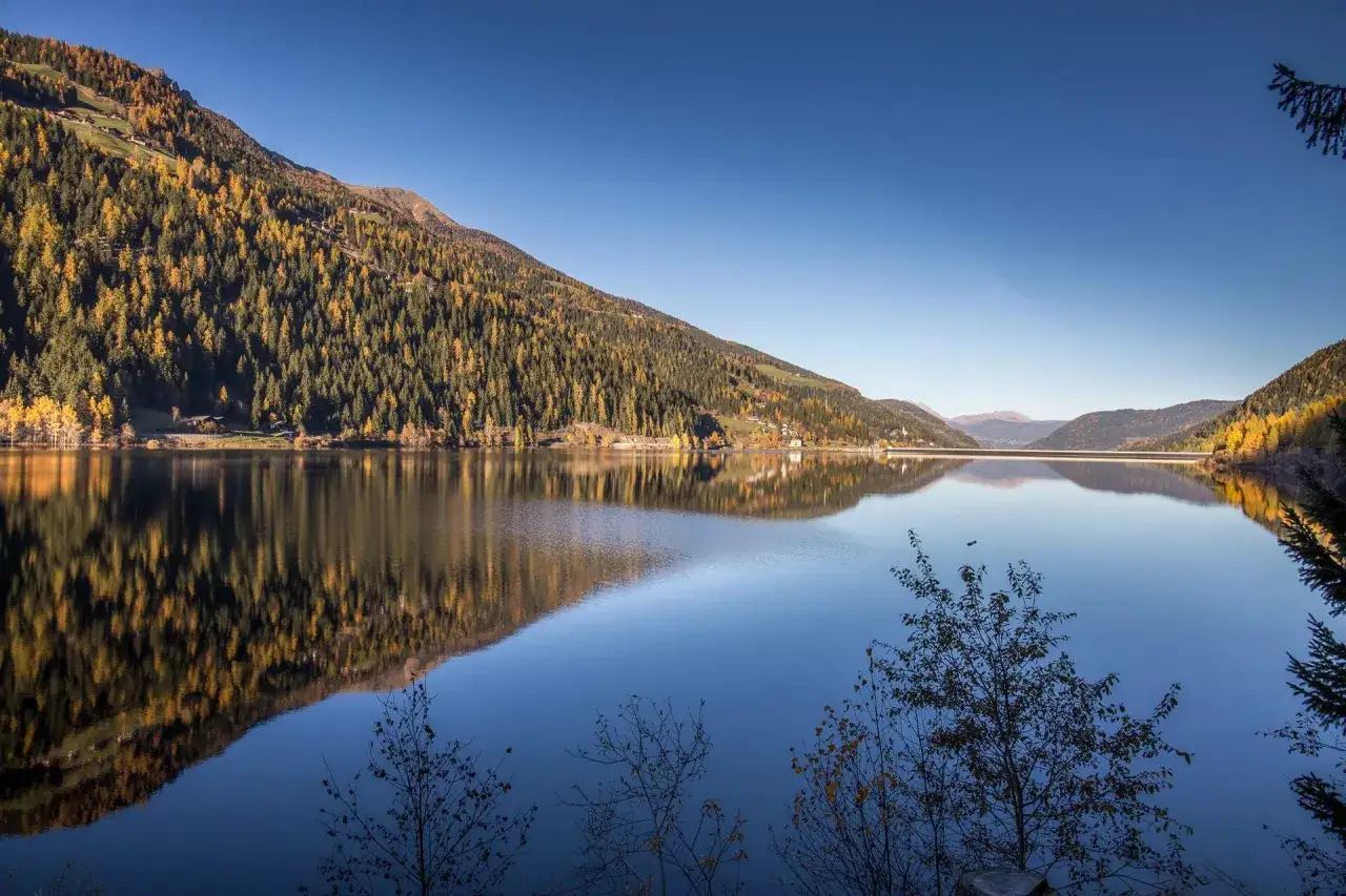 Autunno dorato sul **Val d'Ultimo lago**, con boschi che si specchiano nelle acque calme e un cielo terso.