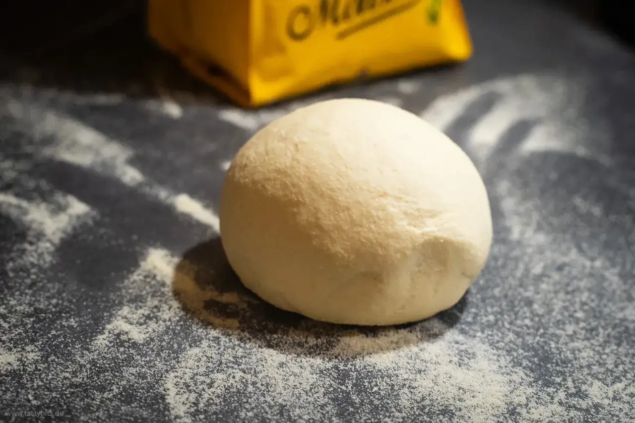Ein runder Ball aus nudelteig ohne ei liegt auf einer bemehlten Arbeitsfl&auml;che. Im Hintergrund ist eine gelbe Packung zu sehen.