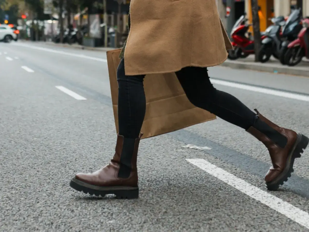 Mujer con botas chelsea marrones y abrigo camel cruza la calle, llevando una bolsa de compras. Un outfit perfecto para el d&iacute;a a d&iacute;a.