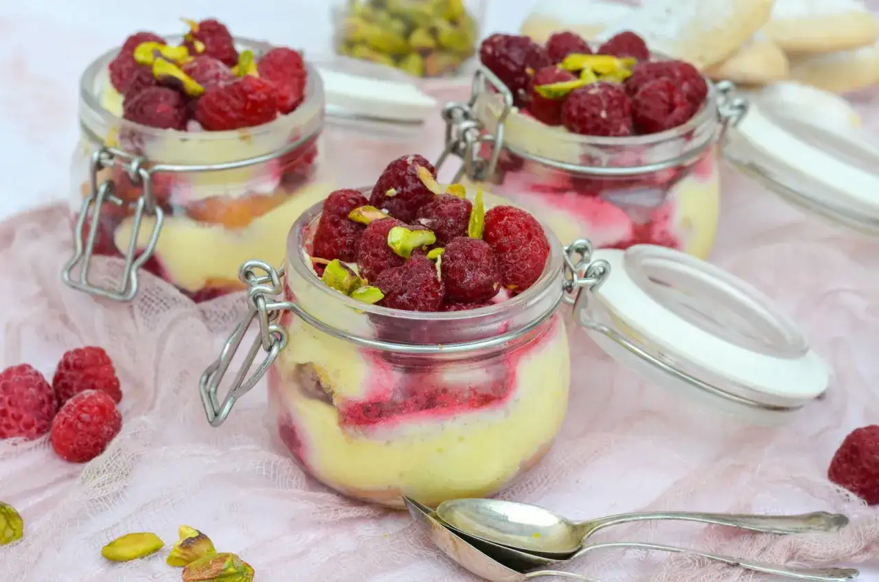 Trois pots de panna cotta framboise, garnis de fruits rouges frais et de pistaches concass&eacute;es, sur un tissu rose.