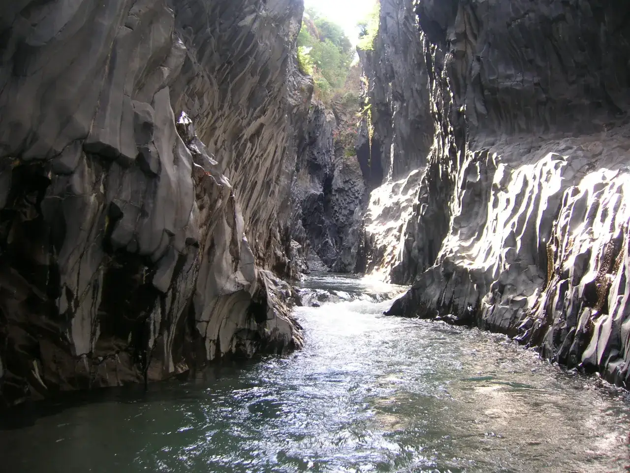 Fiume scorre tra imponenti pareti rocciose scolpite nella **Valle dell'Alcantara**. La luce solare illumina le rocce creando un gioco di ombre e riflessi sull'acqua.