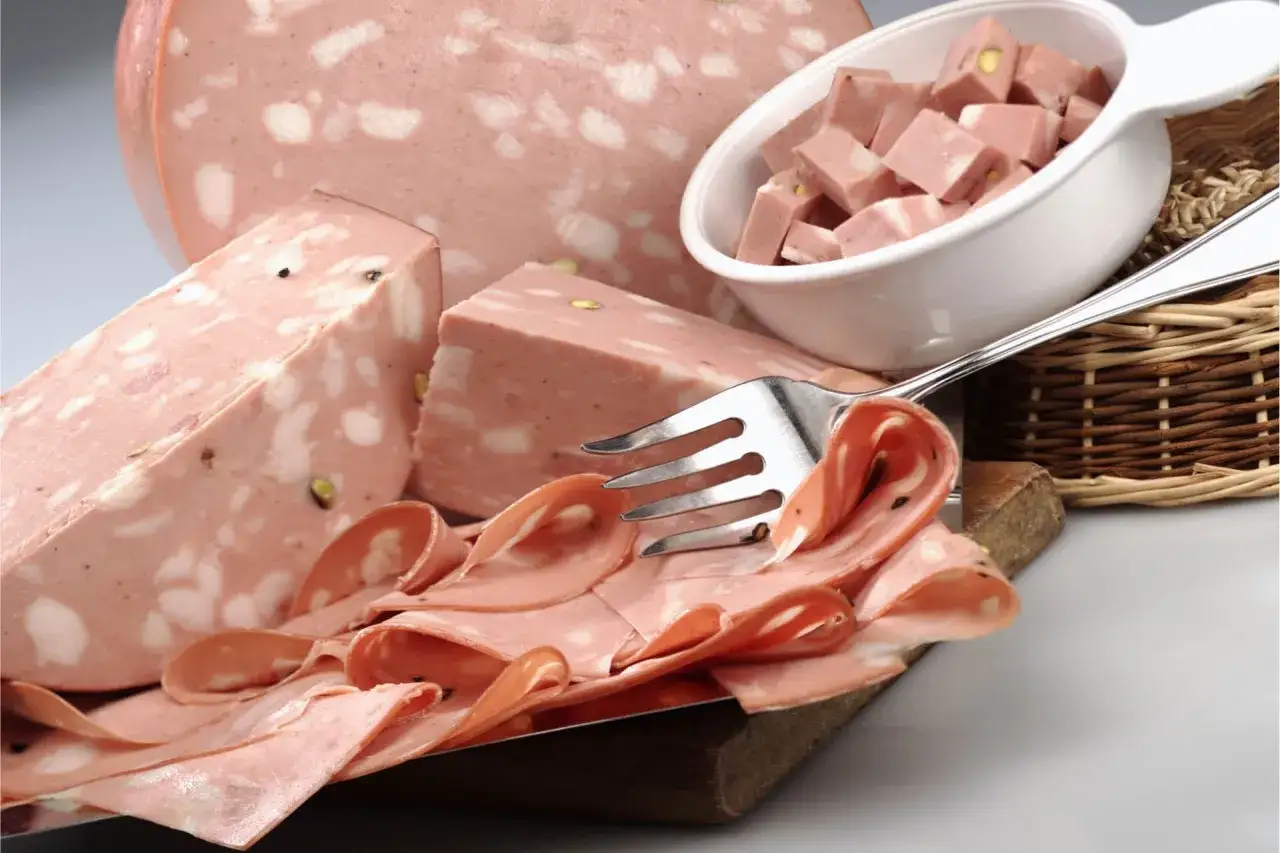 Fetta di mortadella con pistacchi e pepe, servita su un tagliere con fette arrotolate e cubetti in una ciotola.