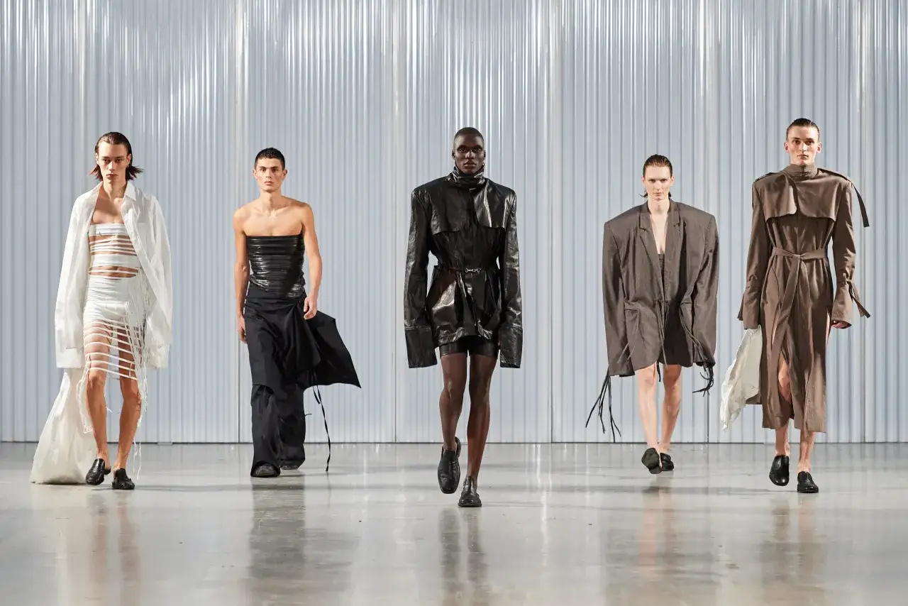 Cinco modelos desfilan con atuendos vanguardistas. Un modelo andrógino luce un traje blanco con flecos, otro un top negro y falda.
