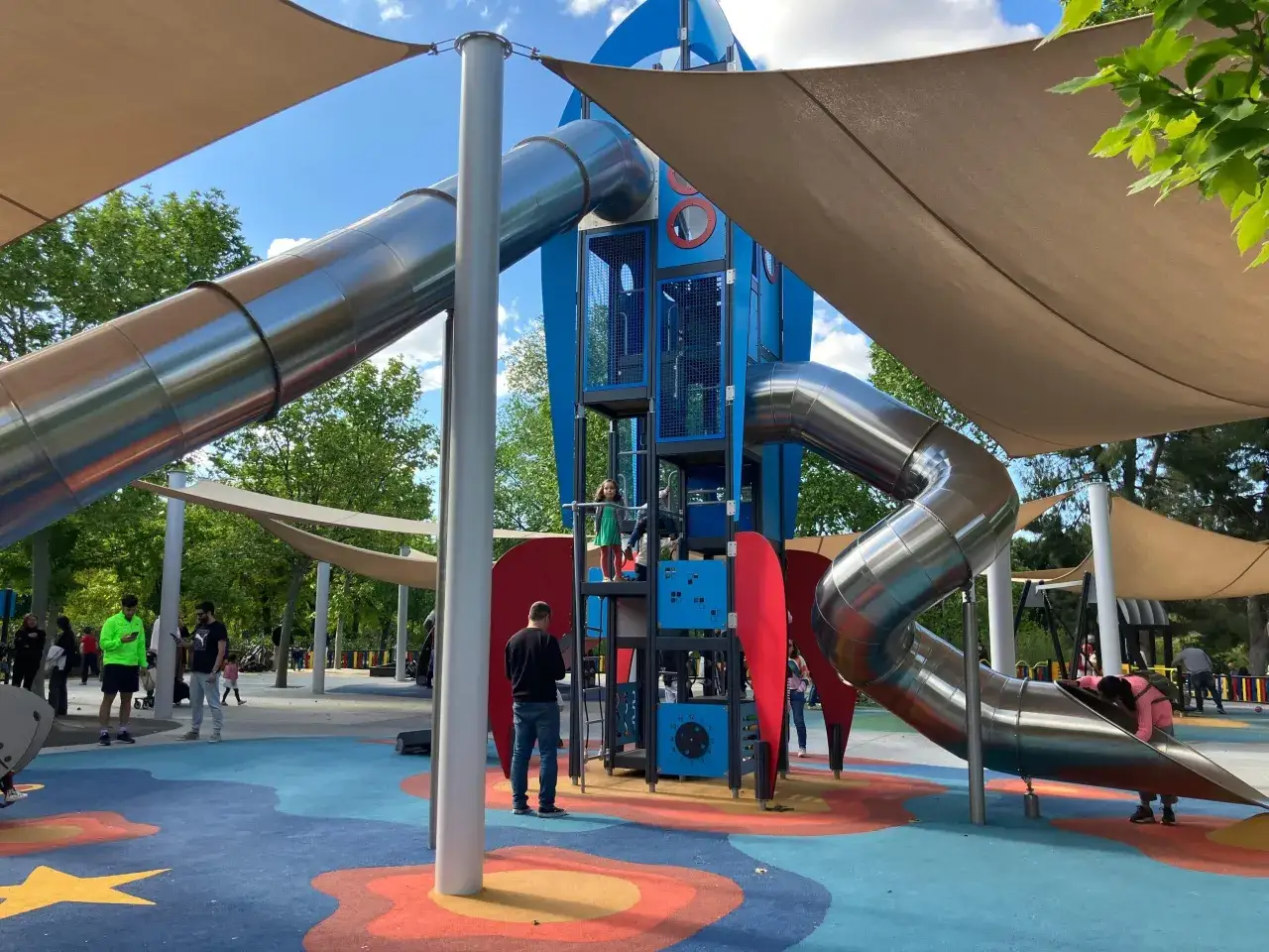 Un colorido parque infantil en Madrid con toboganes met&aacute;licos en forma de cohete y toldos para proteger del sol.