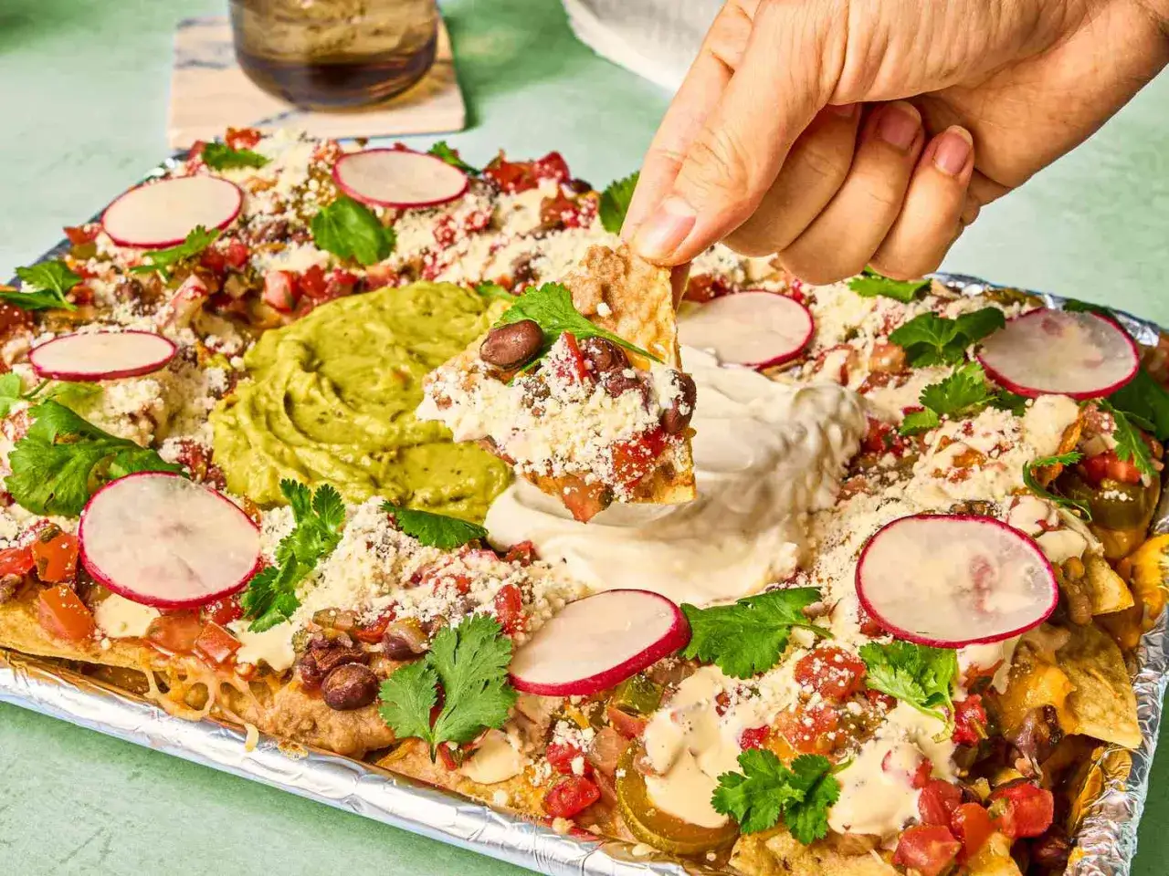 Eine Hand greift nach einem Chip von einem riesigen, beladenen **Nacho Salat** mit Guacamole, Sauerrahm, Bohnen, Tomaten und Radieschen.