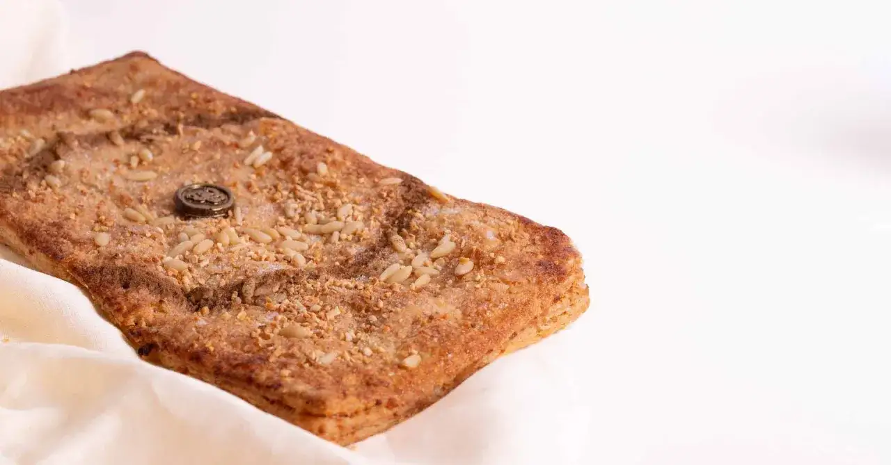 Deliciosa coca de llardons con pi&ntilde;ones y un toque de az&uacute;car, sobre un pa&ntilde;o blanco.