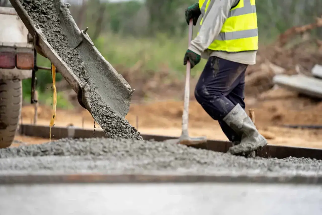 Betoniarka wylewa świeży beton, pracownik w kamaszach i kamizelce odblaskowej go rozprowadza. Ważne jest, jak długo podlewać beton, by był trwały.