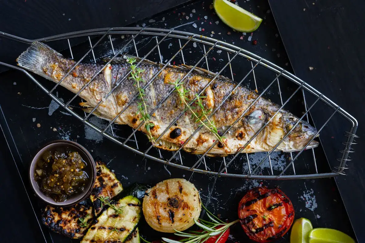 Ein ganzer Wolfsbarsch vom Grill, garniert mit Kräutern, daneben gegrilltes Gemüse, Tomaten und eine Limettenspalte.