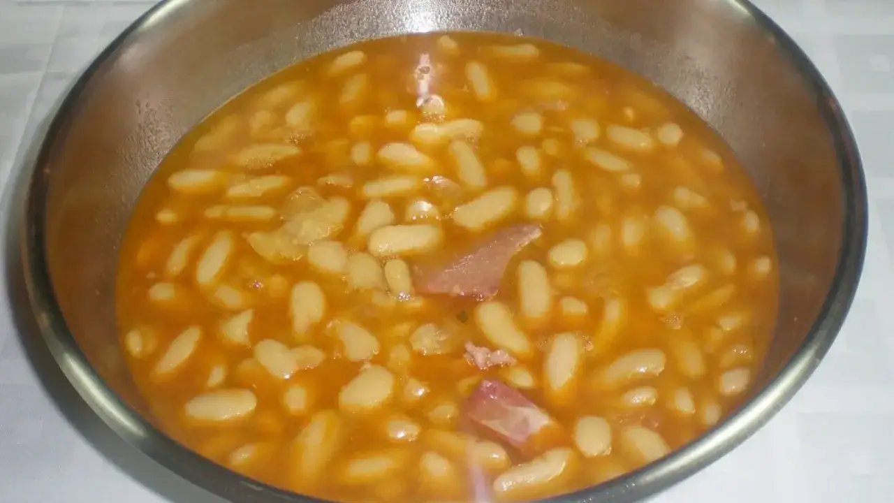 Un cuenco metálico lleno de alubias blancas en un caldo anaranjado con trozos de jamón.