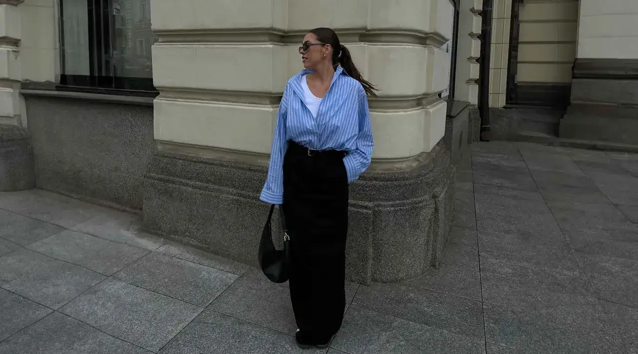 Une femme porte une chemise rayée bleue et blanche, une jupe longue noire et un sac à main. Ce look avec marinière femme est chic et décontracté.