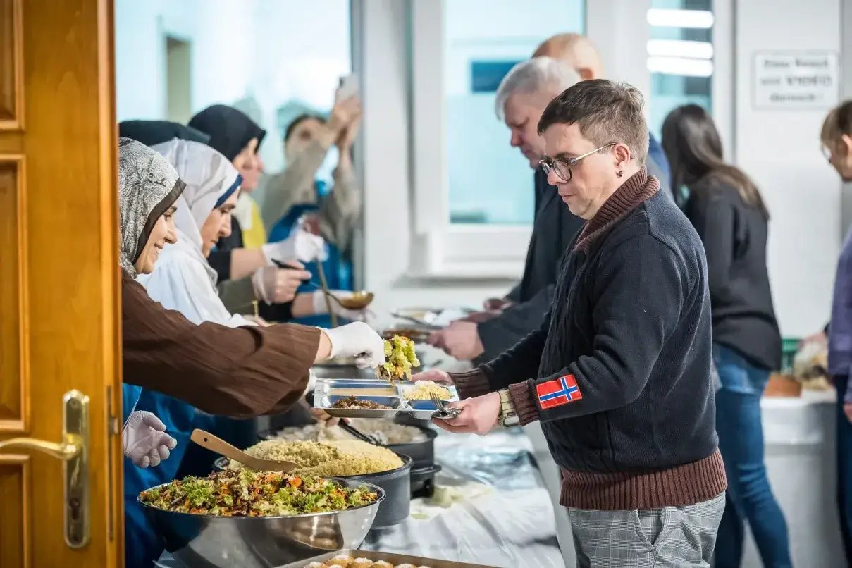 Menschen holen sich Essen bei einem Festmahl zum Eid. Eine Frau mit Kopftuch bedient einen Mann mit Brille.