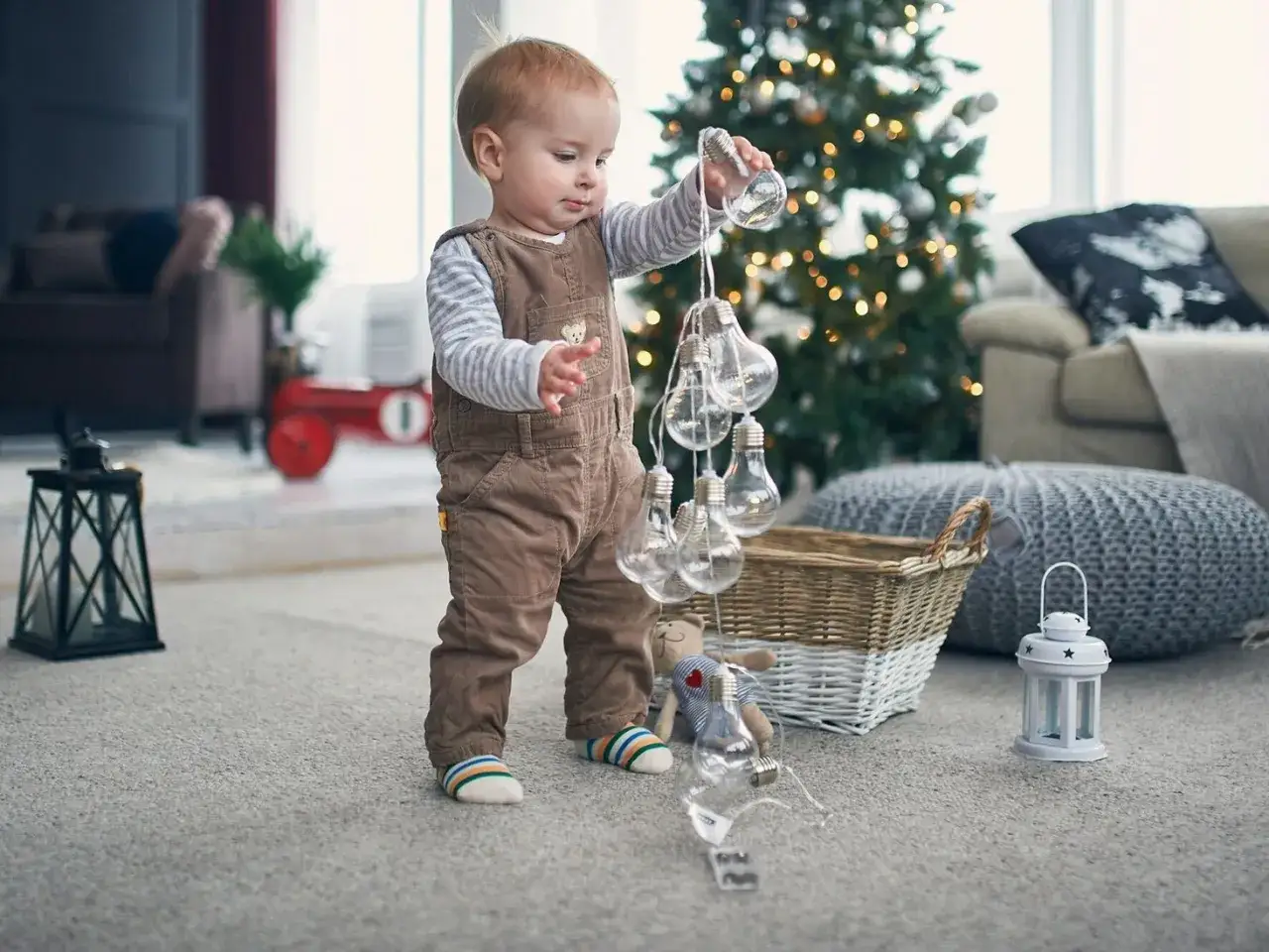 11 miesięczne dziecko w brązowych ogrodniczkach bawi się lampkami choinkowymi. W tle choinka i kanapa.