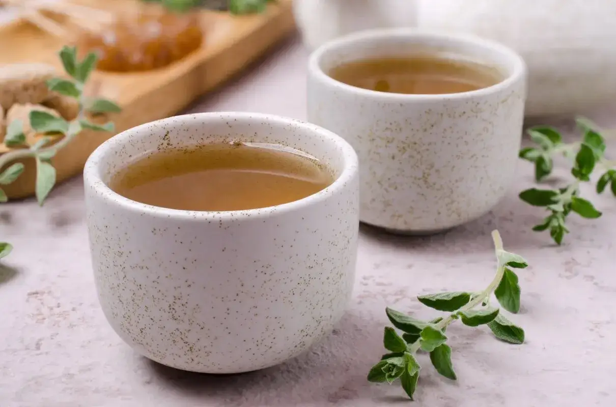Dwie filiżanki z herbatą z oregano, kt&oacute;ra pomaga na przeziębienie i wspiera trawienie. Obok gałązki świeżego oregano.