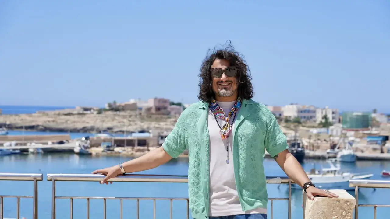 Uomo con occhiali da sole e maglia verde sorride con il porto di Lampedusa alle spalle, scenario di
