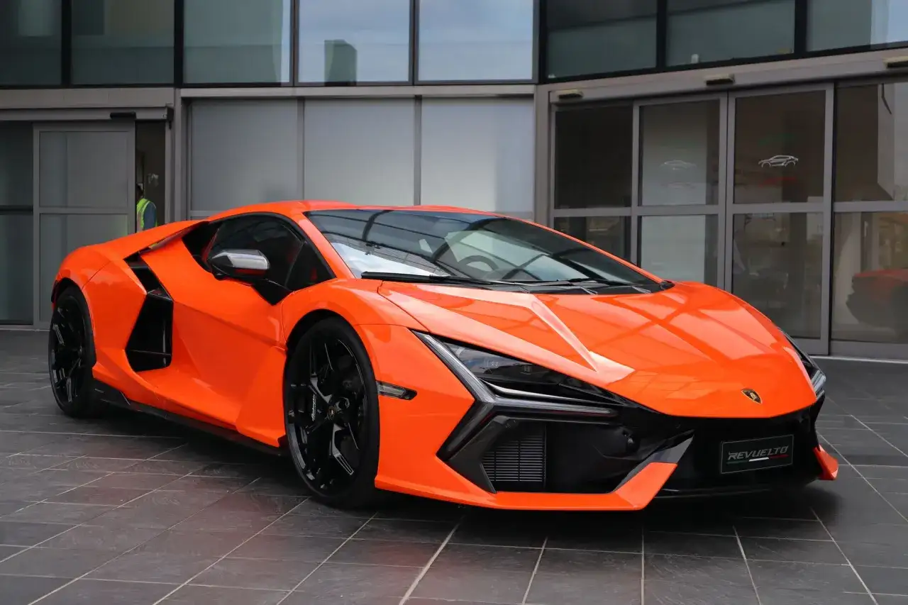 Pomarańczowy Lamborghini Revuelto z czarnymi felgami stoi przed nowoczesnym budynkiem.