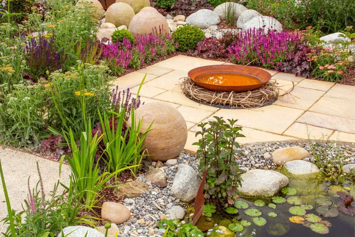 Ein blühender garten ohne rasen mit Teich, Feuerstelle und Steinkugeln. Bunte Stauden und Gräser schaffen ein lebendiges Bild.