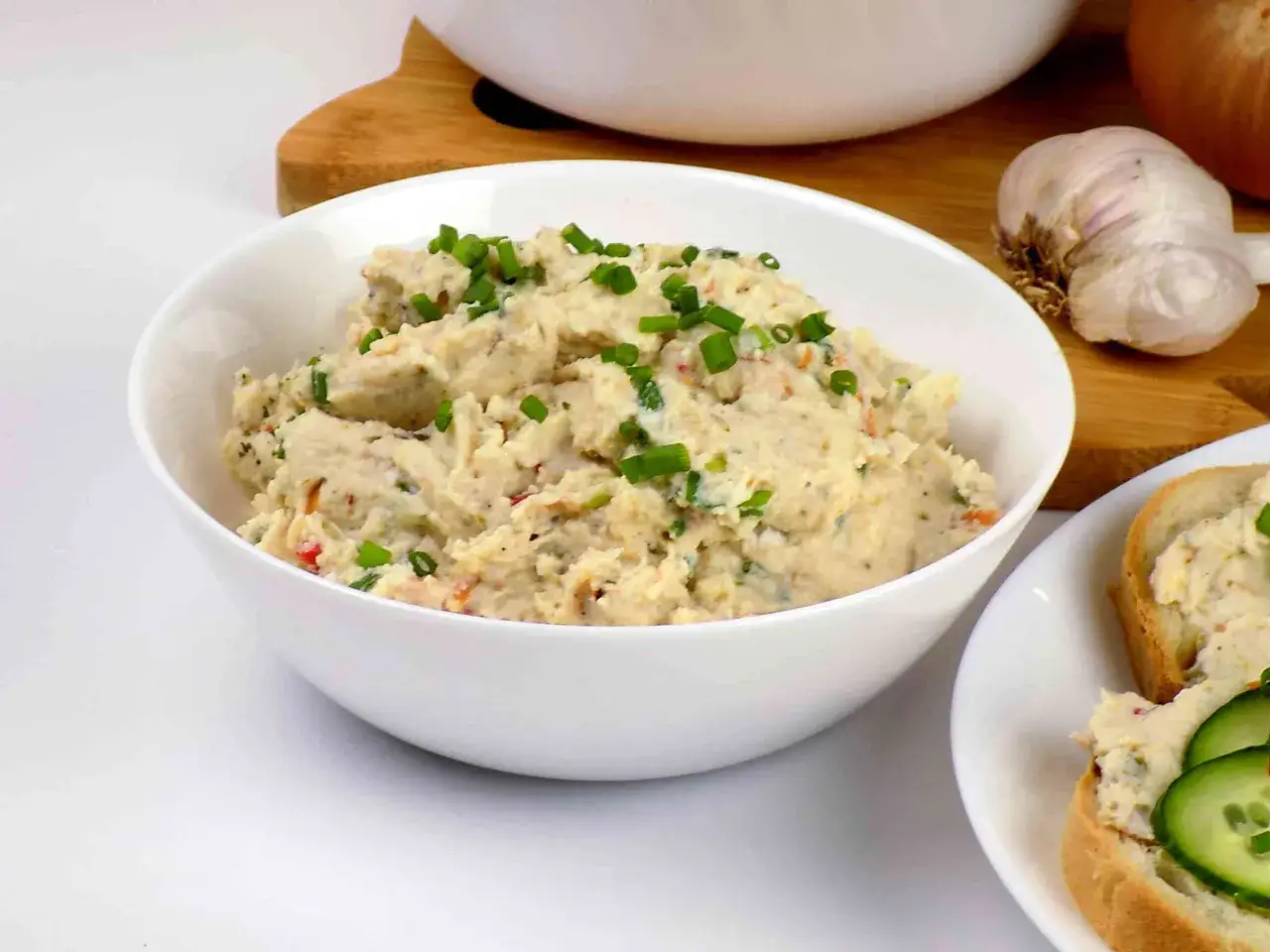 Pyszna pasta z kurczaka, posypana szczypiorkiem, w misce. Obok kromka chleba z pastą i og&oacute;rkiem.