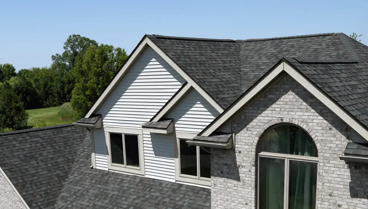 A house roof showcases various colors of asphalt shingles, from dark charcoal to lighter grays, blending with the surrounding trees.