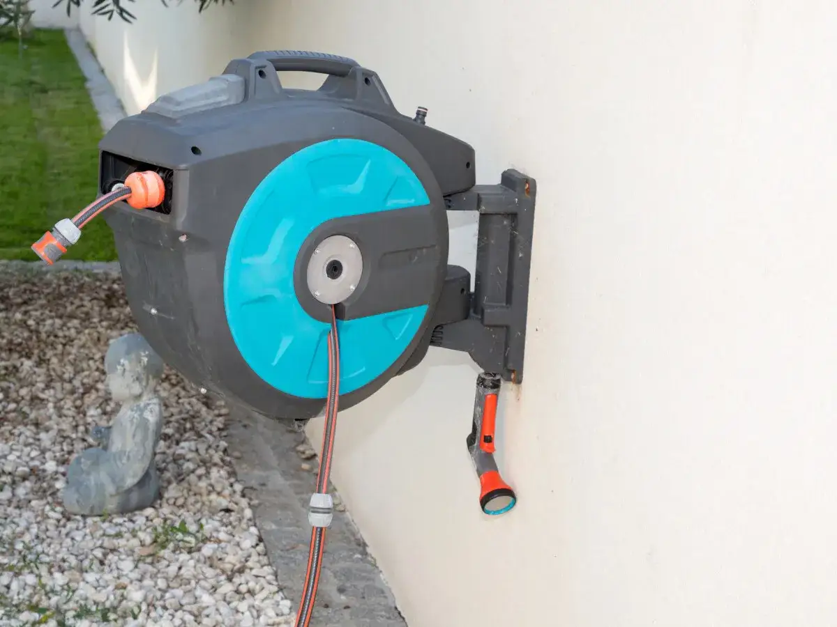 Ein schwarzer wandschlauchaufroller mit türkisfarbenen Akzenten ist an einer weißen Wand montiert. Ein Gartenschlauch mit Sprühkopf hängt daran.