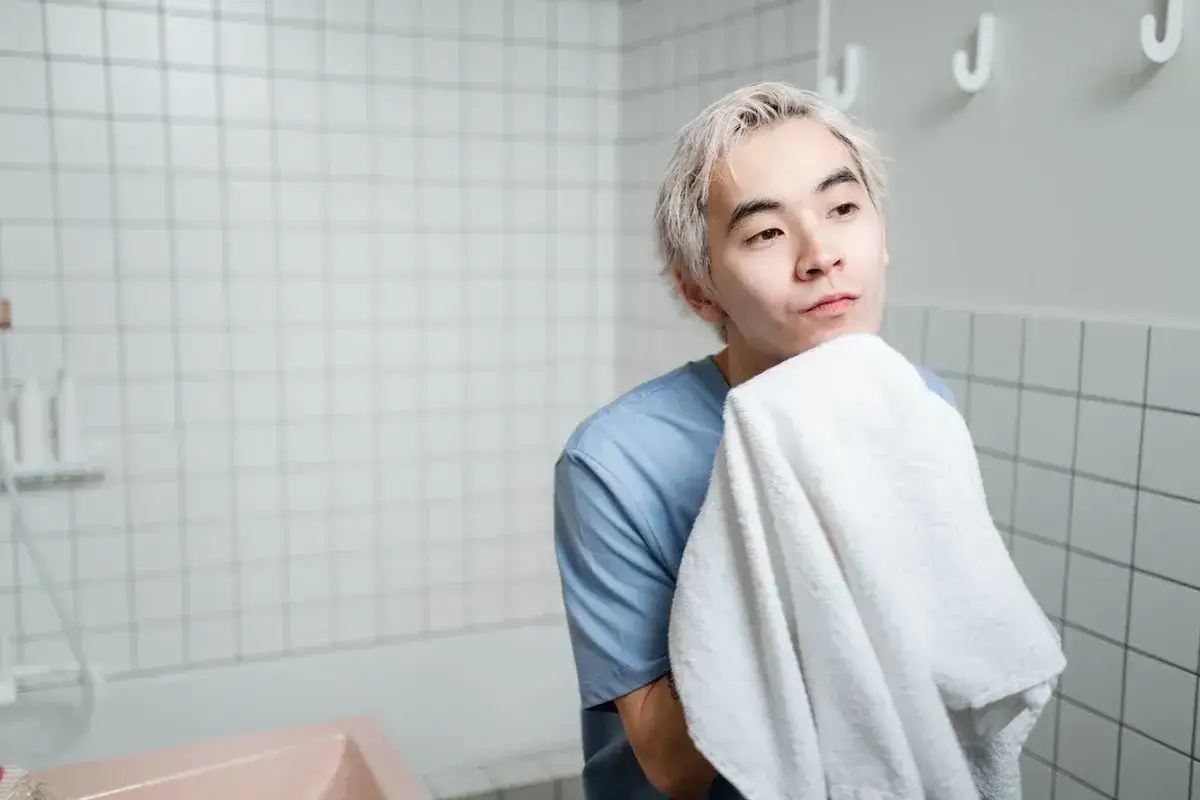 En ung man med blont h&aring;r torkar sitt ansikte med en vit handduk. Han har en bl&aring; tr&ouml;ja p&aring; sig och st&aring;r i ett badrum med kaklade v&auml;ggar. Hans hud ser lite fet ut.