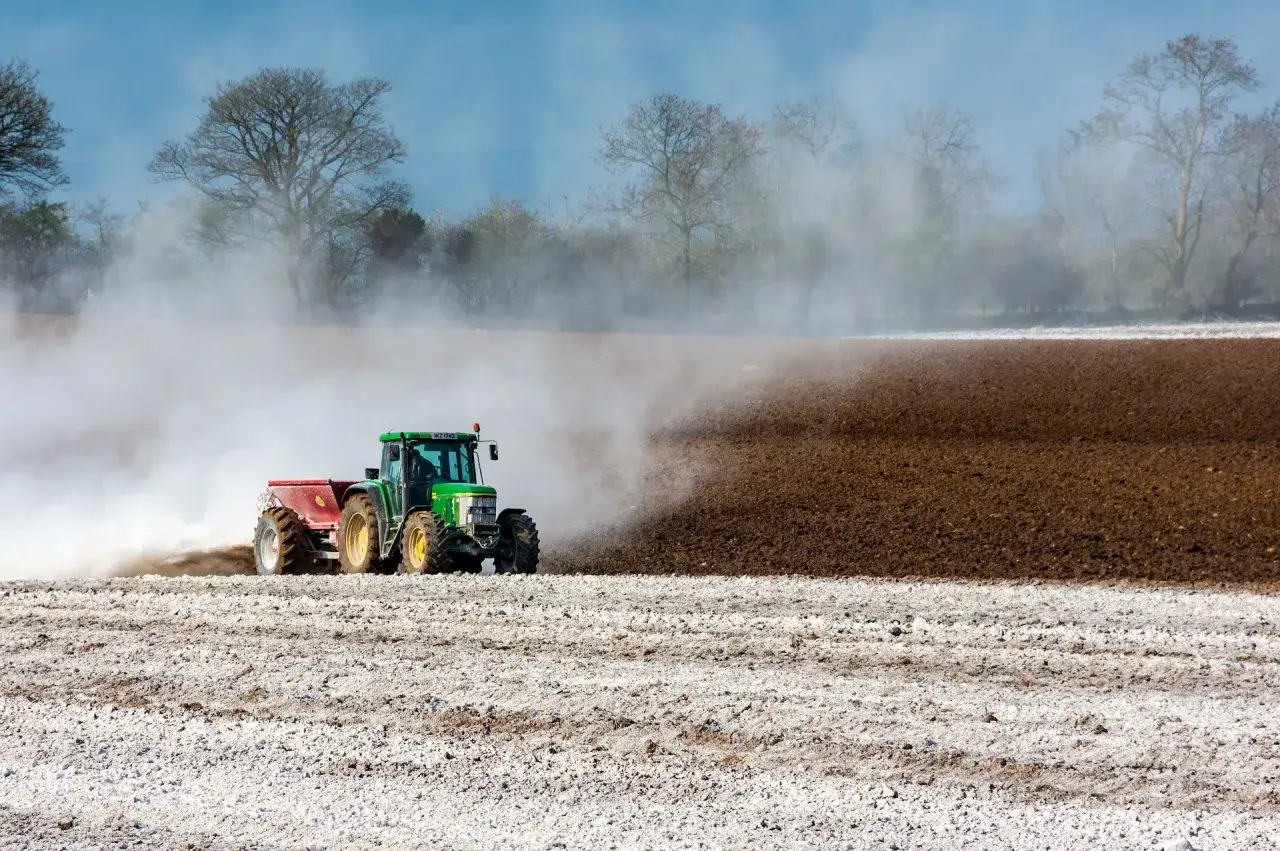 Zielony traktor rozsypuje wapno na polu, tworząc białą smugę na tle oranej ziemi.