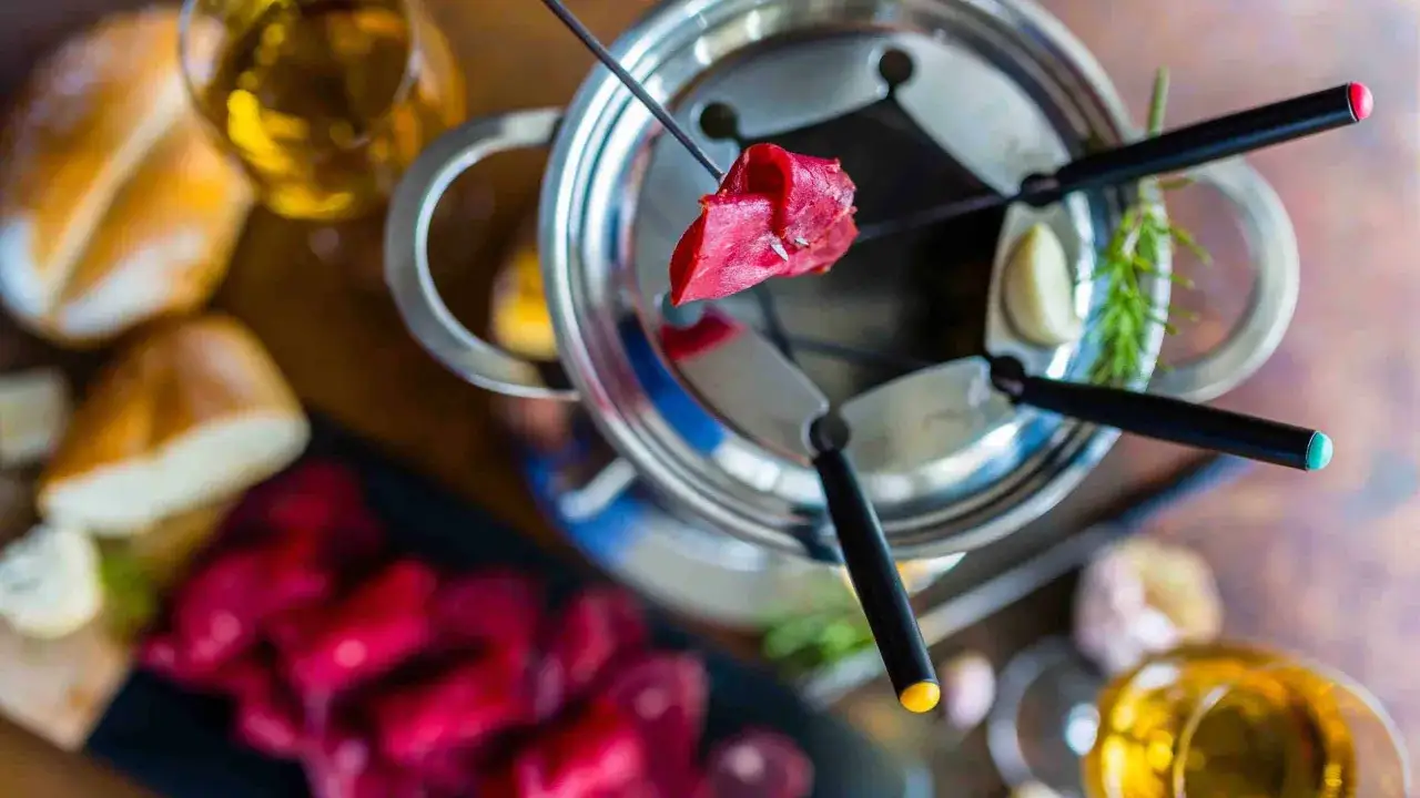 Un morceau de viande rouge est piqu&eacute; sur une brochette, pr&ecirc;t &agrave; &ecirc;tre plong&eacute; dans le bouillon de la fondue chinoise. Autour, du pain, des l&eacute;gumes et des verres de vin.