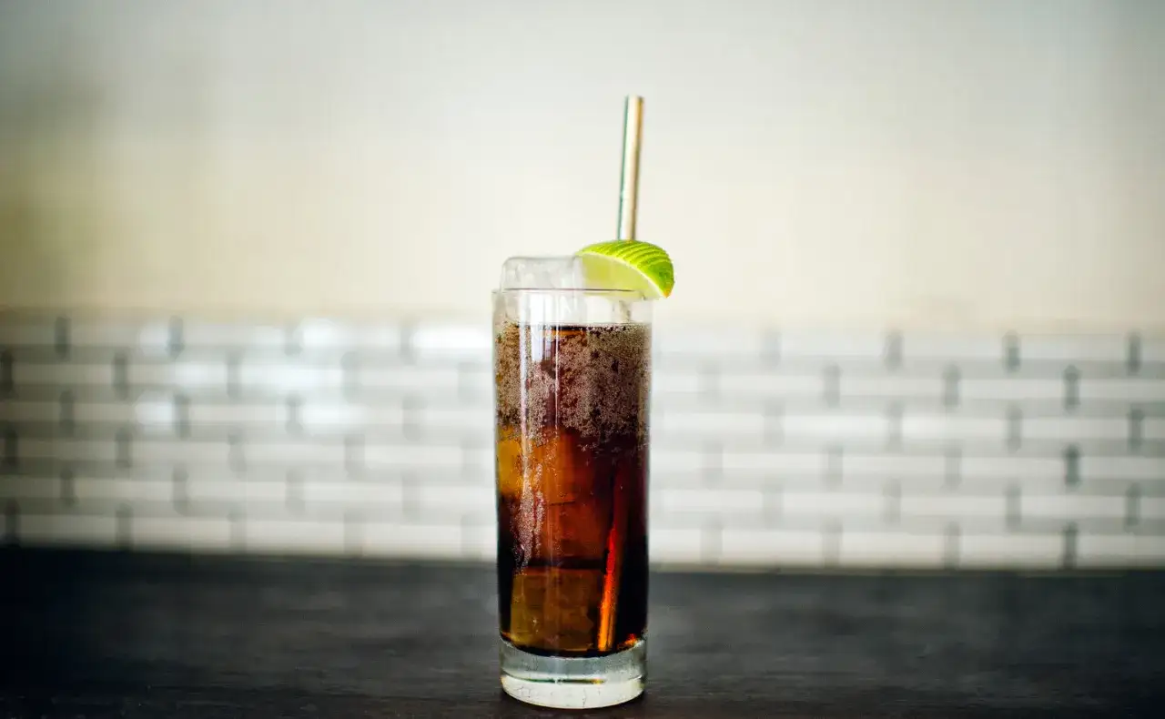 Idealny Cuba Libre przepis: wysoka szklanka z lodem, ciemnym napojem, plasterkiem limonki i słomką. Orzeźwiający drink na upalne dni.