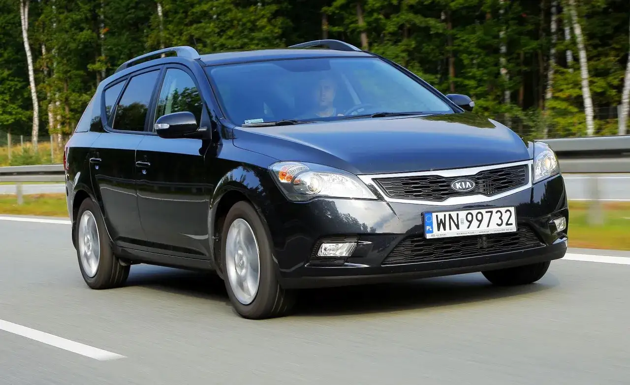 Czarny Kia Ceed 1. kombi jedzie po drodze, otoczony zielenią drzew.