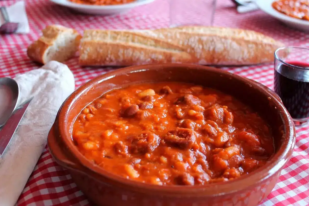 Un cuenco de barro con alubias con chorizo, pan rústico y una copa de vino tinto.