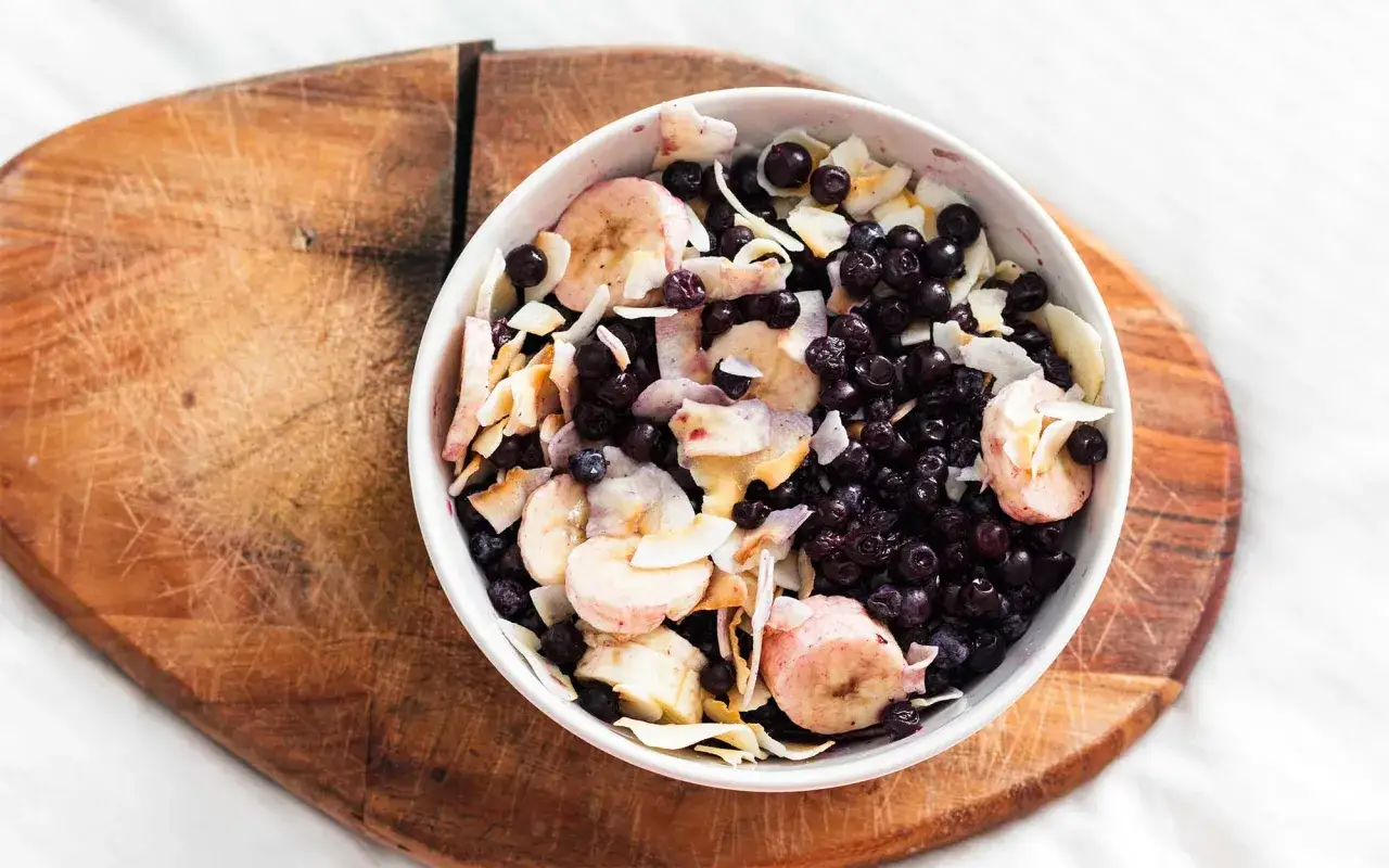 Eine k&ouml;stliche vegane Smoothie Bowl mit Blaubeeren, Bananenscheiben und Kokoschips auf einem Holzbrett.