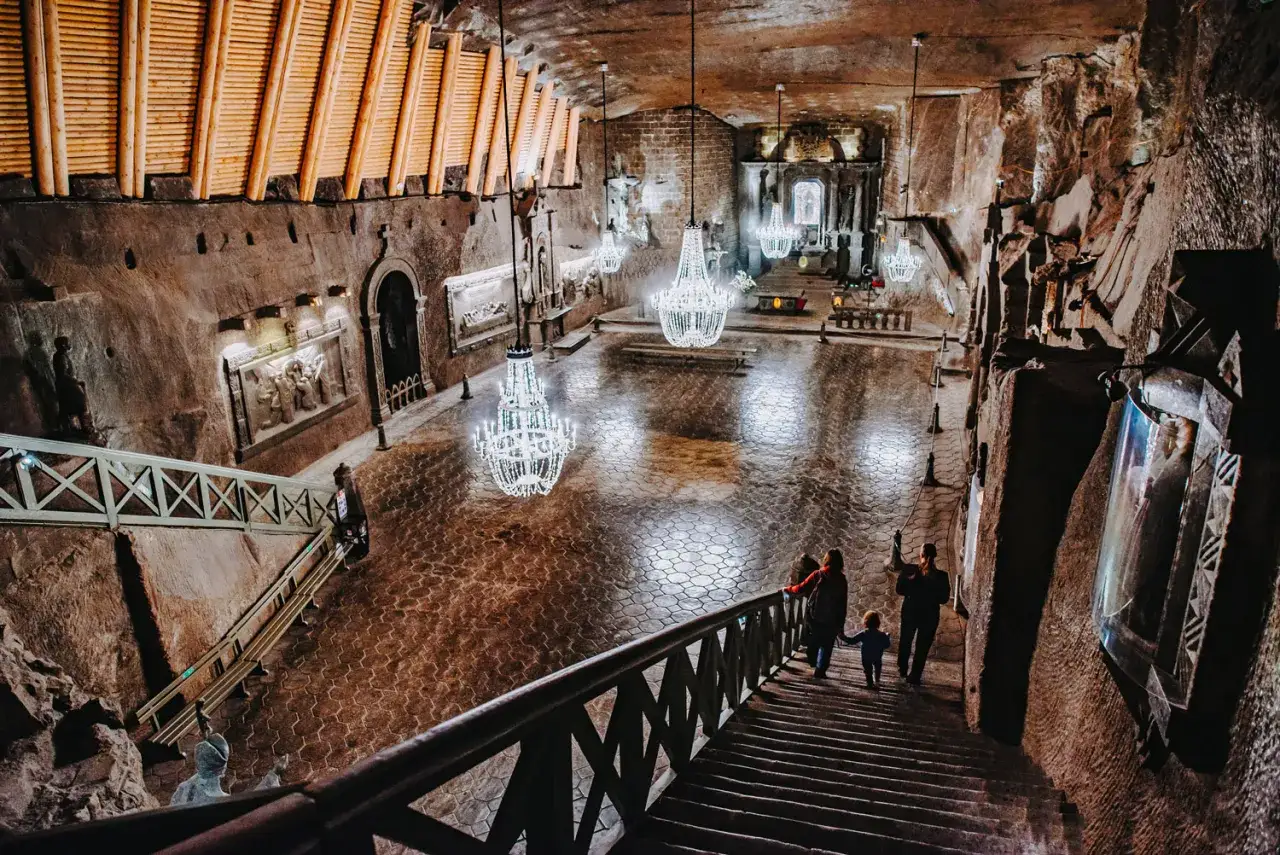 Rodzina schodzi po schodach do kopalni soli w Wieliczce. W podziemnej komorze wiszą kryształowe żyrandole, a ściany zdobią solne rzeźby.