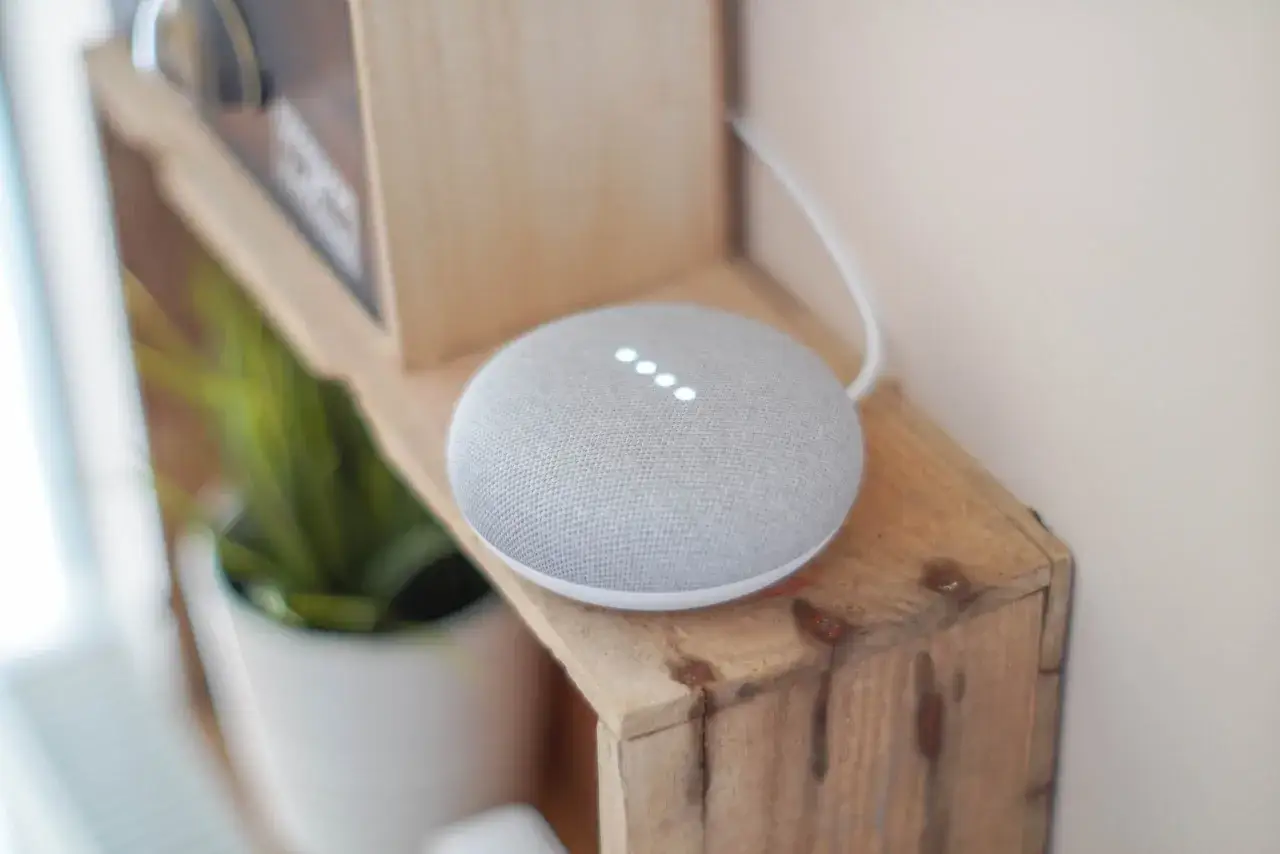 Grauer Smart Speaker mit leuchtenden Punkten, der auf einem Holzregal steht. Er wartet auf einen Befehl, der durch Voice Match erkannt wird.