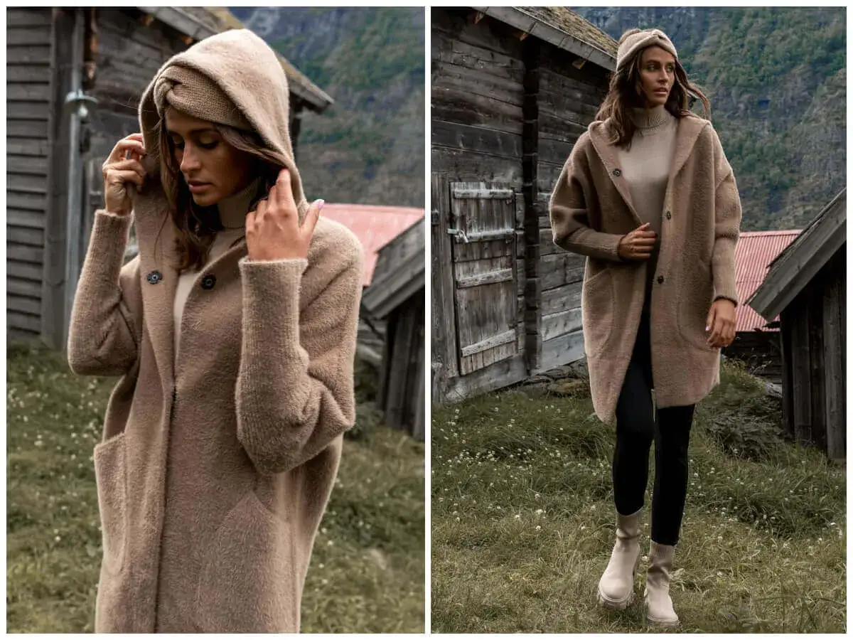 Jass płaszcze z alpaki: modelka w beżowym, miękkim płaszczu z kapturem, otoczona rustykalną scenerią górskich chat.