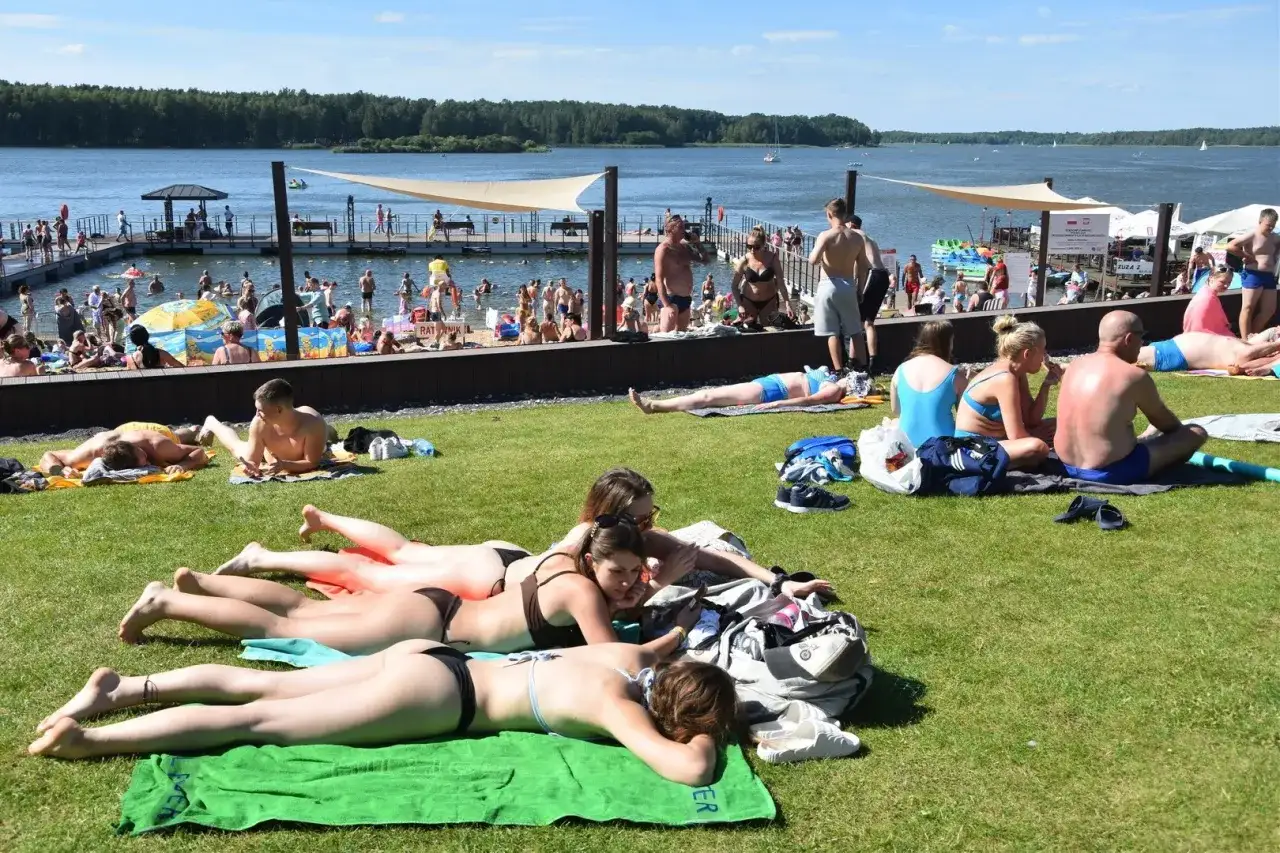 Ludzie opalają się na trawie nad **zalewem koronowskim**. W tle widać wodę, pomost i plażowicz&oacute;w.