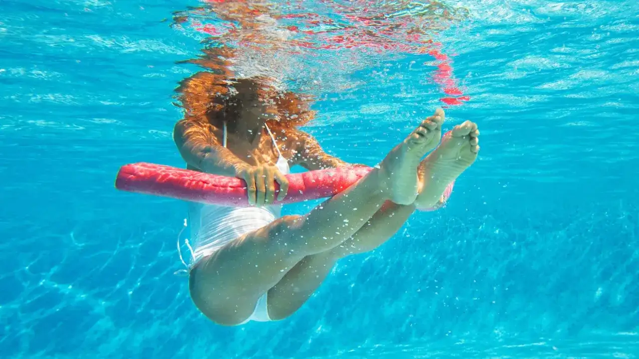 Femme aux cheveux roux faisant un exercice piscine, tenant une nouille de piscine rose sous l'eau.