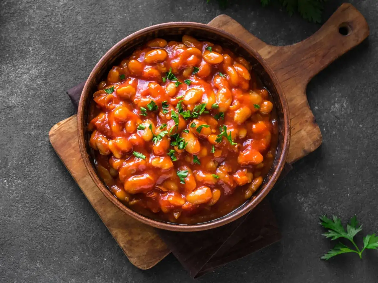 Schmackhaftes baked beans rezept in einer Sch&uuml;ssel, garniert mit Petersilie, auf einem Holzbrett.