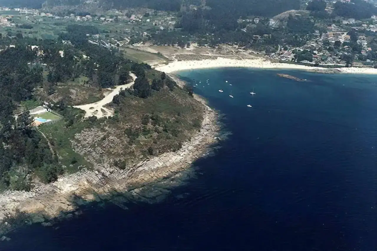Vista aérea de la playa de Limens, con arena blanca, aguas azules y barcos navegando. Al fondo, un pueblo costero y colinas verdes.