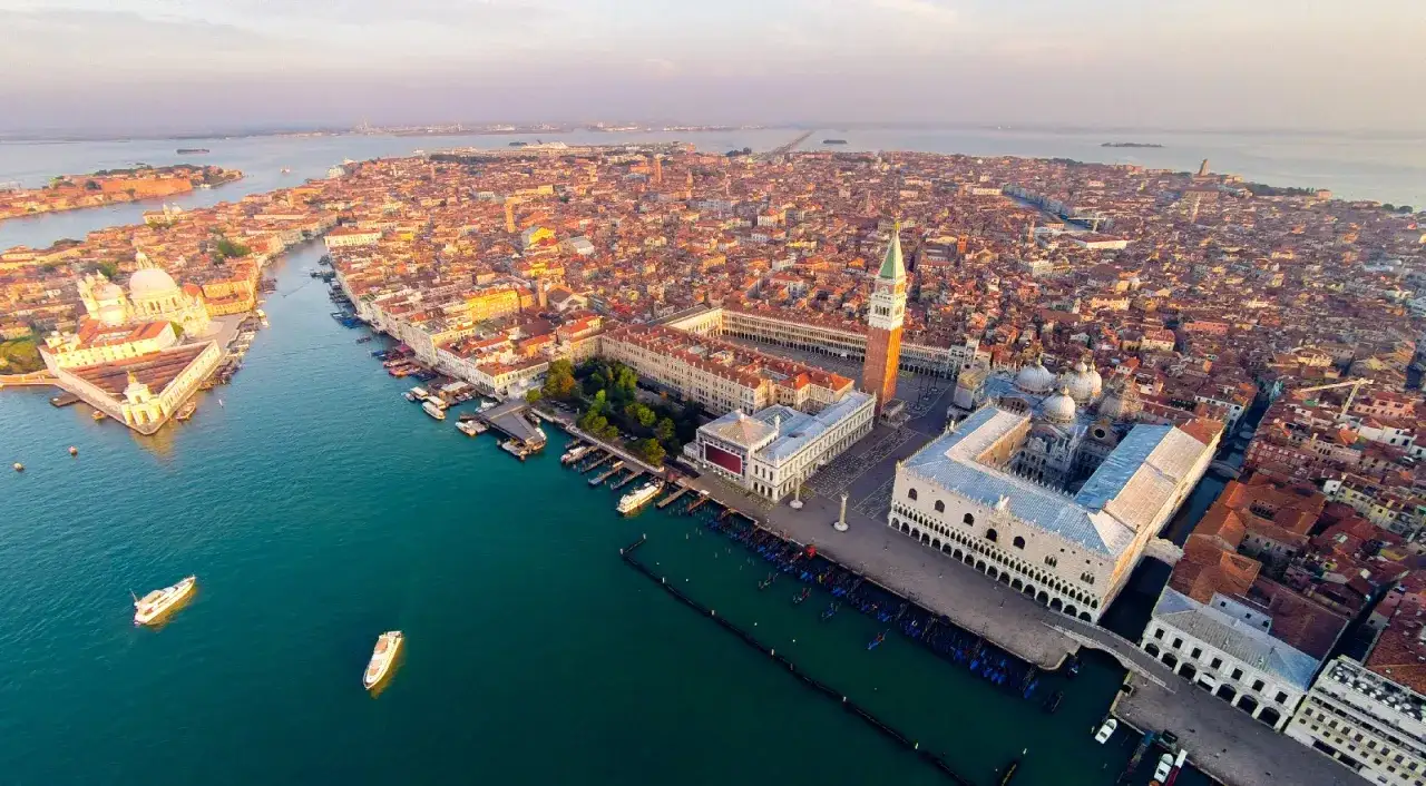 Vista aerea di **Piazza San Marco a Venezia** al tramonto, con il Canal Grande e i tetti rossi della città.